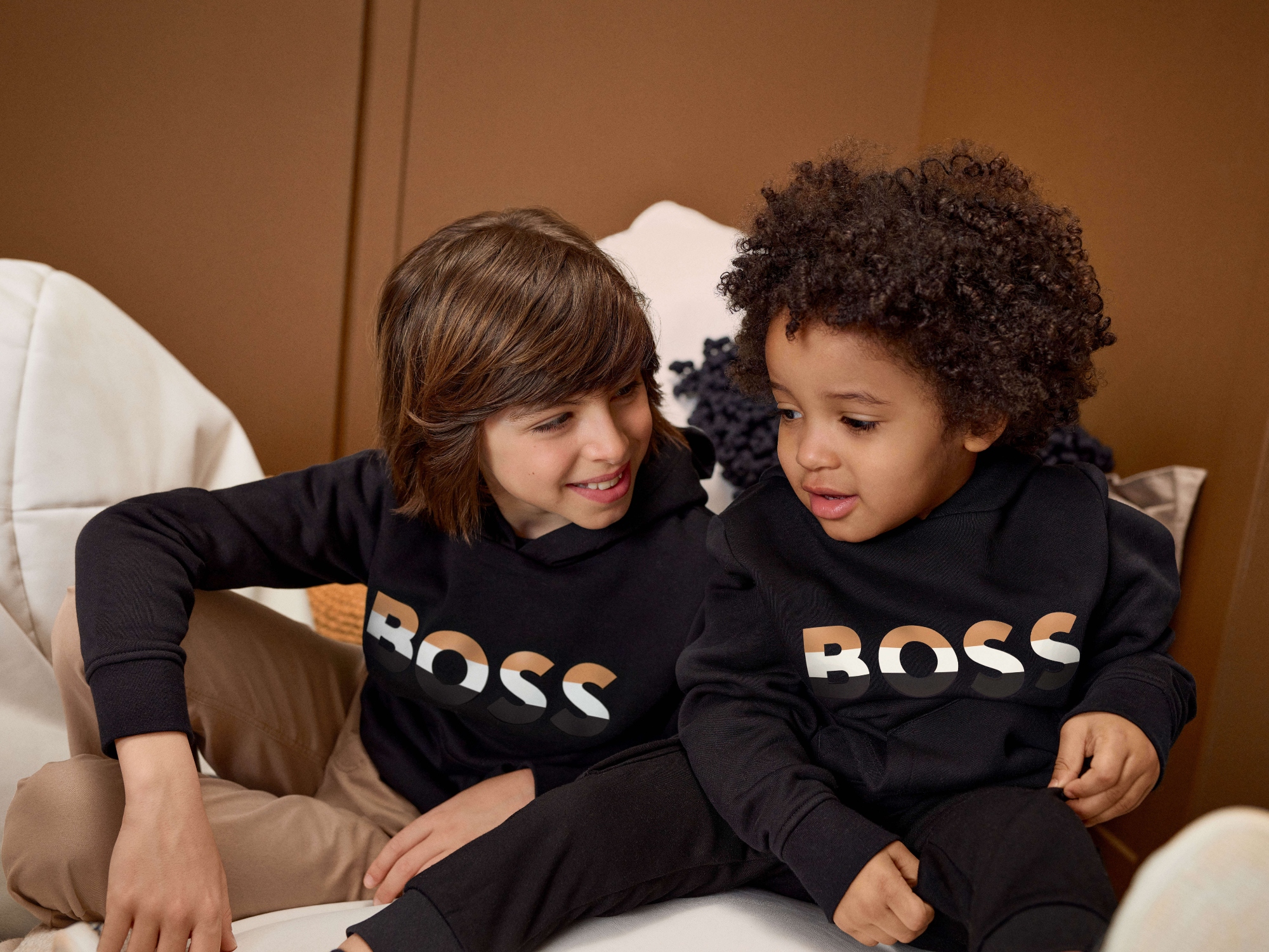 Two kids sat on a bed dressed in Boss